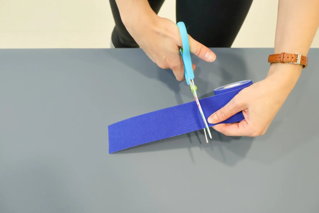 Die Physiotherapeutin, schneidet mit einer Schäre die rechte Ecke eines blauen Tapes rund.
